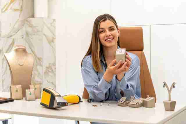 Woman smiling while holding jewellery box at Gold Exchange Universe. Display of gold and gemstone jewellery including earrings and necklace on table. Luxury jewellery shop setting with marble walls.